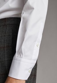 Wit overhemd met lange mouwen en knopen, gedragen met donkergrijze geruite broek op een effen grijze achtergrond.