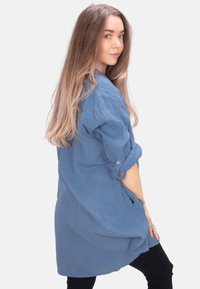 Blaue, strukturierte Tunika-Bluse mit hochgerollten Ärmeln, Knopfakzenten und Seitentaschen, kombiniert mit schwarzen Hosen.