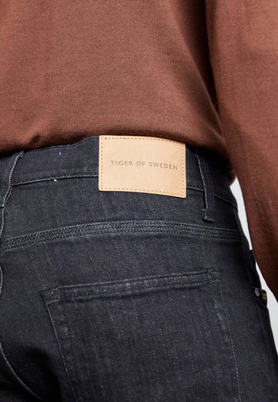 Dark denim jeans with a leather patch labeled "TIGER OF SWEDEN," showing intricate stitching and a smooth texture. Brown shirt visible above.