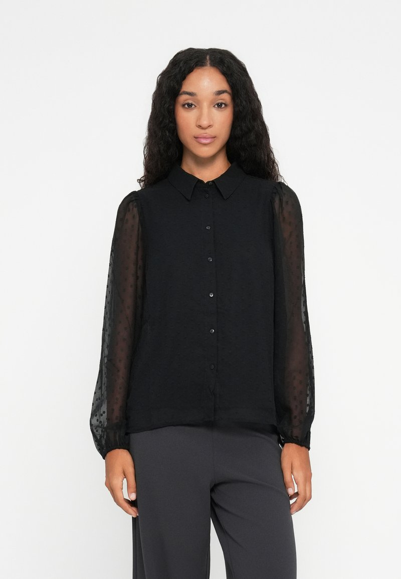 Blouse noire avec un col, une fermeture à boutons sur le devant et des manches bouffantes transparentes. Le tissu présente une texture à pois subtils.