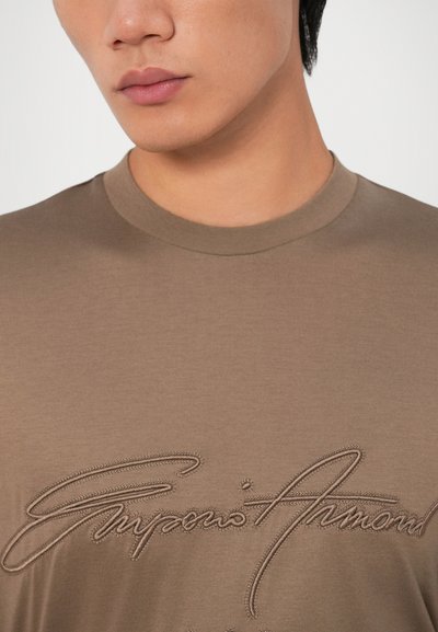 Camiseta de algodón marrón con escote redondo, que presenta un logotipo bordado prominente en un tono más claro de marrón en el pecho.