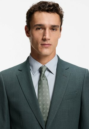 Completo verde scuro con trama strutturata e rever a lancia, indossato sopra una camicia azzurra. La cravatta presenta un motivo geometrico in verde più chiaro e bianco.