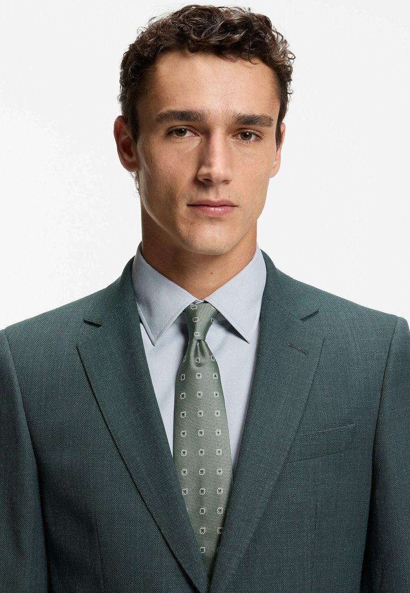 Donkergroen textuurpakk met inkeping revers, gedragen over een lichtblauw overhemd. De stropdas heeft een geometrisch patroon in lichter groen en wit.
