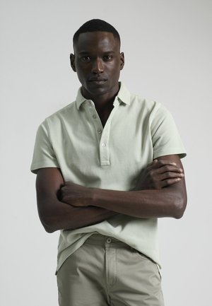 Camisa polo verde claro con acabado texturizado, tapeta de tres botones y mangas cortas, combinada con pantalones beige claros.