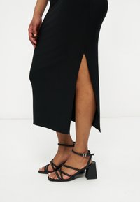 Γυναίκα που φοράει μαύρα σανδάλια με λουριά και τετράγωνο τακούνι, μαζί με μαύρη midi φούστα με πλαϊνή σχισμή, στέκεται μπροστά σε λευκό απλό φόντο.