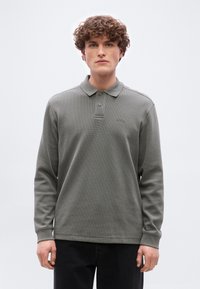 Νεαρός άνδρας με σγουρά μαλλιά, φορώντας γκρι μακρυμάνικη πόλο μπλούζα με δύο κουμπιά και μαύρο παντελόνι, στέκεται μπροστά σε απλό φόντο.