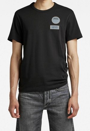 Homme portant un t-shirt noir à manches courtes avec deux petits écussons bleus sur la poitrine et un jean en denim gris, debout sur fond blanc.