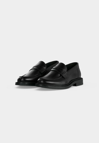 Ζευγάρι μαύρα δερμάτινα loafers με χαμηλά τακούνια και λεπτομέρεια με λουράκι στην πάνω πλευρά, εμφανισμένα σε απλό φωτεινό φόντο.
