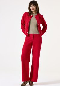Veste rouge texturée avec un pantalon large assorti et un pull beige en laine douce en dessous. Les détails incluent des fermetures à bouton et des poches avant.