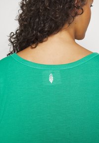 Top acanalado verde con un amplio escote y un pequeño emblema de hoja blanca en la parte posterior. Textura suave y diseño casual.