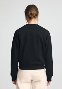 Svart sweatshirt med rund halsringning, sänkta axlar och ribbade ärmslut; bärs med ljusbeige byxor. Slät textur med minimal detaljering.