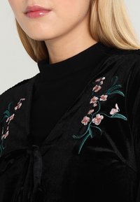 Svart sammetplagg med blommönster i rosa och grönt på framsidan. Har en knytning i midjan och en hög halsringning. Slät textur.