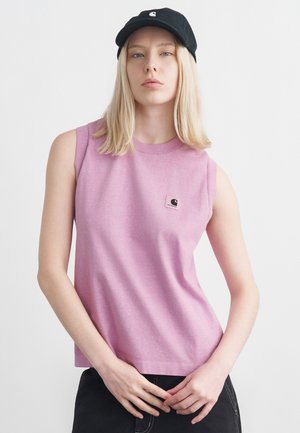 Lyshåret person iført en sort kasket, ærmeløs lilla skjorte med logo og sorte bukser, stående foran en ensfarvet hvid baggrund.