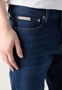 Calvin Klein traperice u tamnoplavoj boji. Značajke uključuju brendiranu etiketu, metalne dugmadi i kontrastnu šav na džepovima.