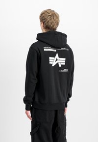 Persoon met kort blond haar, gekleed in een zwarte Alpha Industries hoodie met wit logo en tekst op de achterkant, met de rug naar de camera gekeerd.