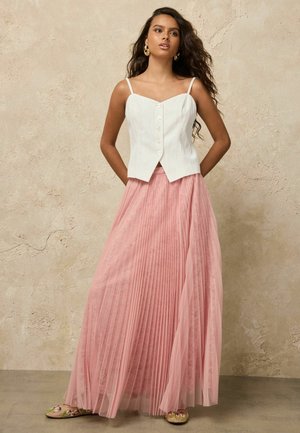 Junge Frau mit langen, welligen Haaren, die goldene Creolen, ein weißes, ärmelloses Oberteil mit Knöpfen und einen rosa plissierten Maxirock trägt, steht vor einer strukturierten Wand.