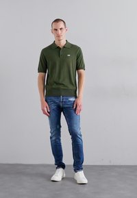 Cămașă polo verde închis cu mâneci scurte, guler cu tiv, și un logo mic, asortată cu blugi decolorati bleu și adidași albi.