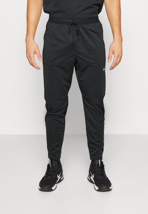 Mann trägt schwarze Nike-Laufhose und schwarze Nike-Sneaker, steht vor einem schlichten hellen Hintergrund.