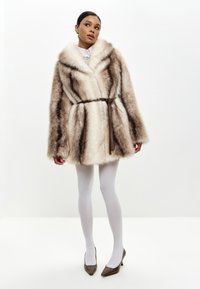 Cappotto in pelliccia sintetica nei toni terra, con un ampio colletto, vita sottolineata da una cintura e maniche lunghe. Modella che indossa collant bianchi e scarpe col tacco marroni.