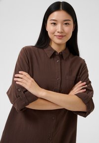 Barna, gombos ing puha anyagból; gallérral, felhajtott ujjakkal és mellzsebbel. A modell keresztbe tett karokkal pózol.