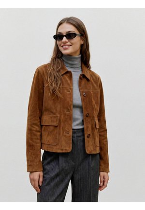 Femme portant une veste en daim marron, un col roulé gris, un pantalon à fines rayures sombre et des lunettes de soleil noires et étroites, souriant légèrement de côté.