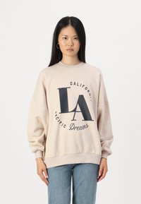Felpa beige con scollo rotondo, spalle scese e testo grafico blu navy che recita "LA California Pacific Dreams." Vestibilità casual, texture morbida.