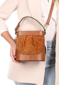 Braune Leder- und Wildleder-Eimer-Tasche mit einer vorderen Faltenoptik, kurzem Griff und abnehmbarem Gurt, ausgestattet mit einer strukturierten Oberfläche und glatten Akzenten.