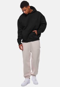 Sort oversized hættetrøje med frontlomme og broderet logo, parret med lysebeige sweatpants med broderet detalje, båret med hvide sneakers.