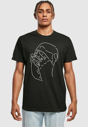 Fekete pamut póló, minimalista fehér vonalas rajzzal, amelyen kezek tartanak egy kerek tárgyat. Rövid ujjú, kerek nyakú design.