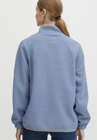 Ljusblå fleecepullover med hög krage, texturerad yta och elastiska ärmslut. Har en rak fåll med en kontrasterande beige kant.