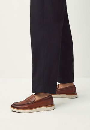 Personne portant un pantalon noir court et des mocassins en cuir marron à enfiler avec des semelles blanches, debout sur un sol blanc.