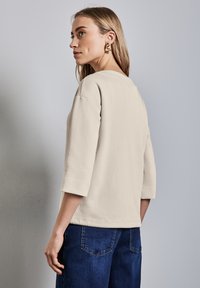 Beige dlouhý rukáv top s kulatým výstřihem a volným střihem. Bavlněný materiál s jemnou texturou. Nosený s modrými denimovými kalhotami.