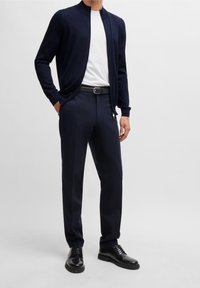 Laivastonsininen neuletakki, valkoinen paita, räätälöidyt laivastonsiniset housut, musta vyö ja mustat nauhalliset kengät. Sileä kangas, kapealahkeiset ja minimalistinen muotoilu.