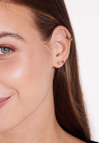 Boucles d'oreilles dorées avec un clou orné d'un gemme transparent, et un design en forme de cœur, se détachant sur un teint de peau lisse et de longs cheveux bruns.