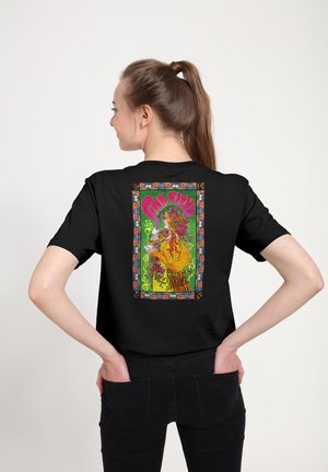 Młoda kobieta z brązowym kucykiem nosi czarny t-shirt z kolorowym, vintage plakatem koncertowym Pink Floyd z kwiatową ramką na plecach.