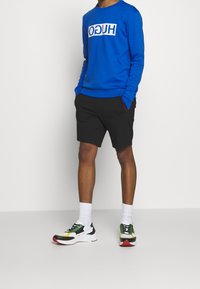 Sweat-shirt bleu à manches longues avec le logo blanc « HUGO », short noir, chaussettes blanches, et baskets multicolores avec des accents verts, jaunes et rouges.