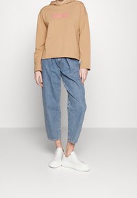 Persona che indossa una felpa beige con il testo "BOSS", jeans blu larghi e sneakers bianche con platform su uno sfondo bianco semplice.