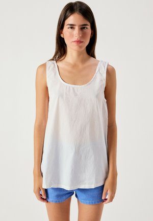 Giovane donna con capelli castani lisci, indossa un top bianco senza maniche e shorts di jeans blu, in piedi davanti a uno sfondo chiaro e uniforme.