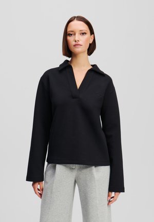 Black, long-sleeve pullover with a collar, featuring a deep V-neck and a subtle logo accent. Soft texture, relaxed fit, paired with gray trousers.