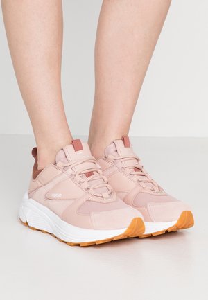 Pari vaaleanpunaisia nauhallisia tennareita, joissa on valkoiset välipohjat ja oranssit pohjat, jalassa paljaina seisomassa sileällä pinnalla.