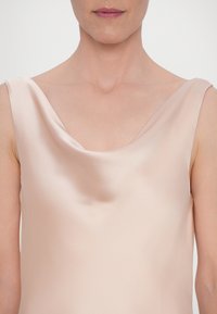 Femme portant un haut sans manches en satin rose pâle avec un col drapé sur un fond clair uni.