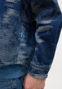 Denimjack in een verloop van blauwtinten met textuurpatronen. Heeft een blauwe "Hugo" label en contrasterende lichte denim accenten.