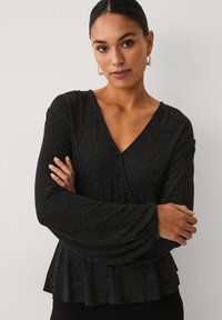 Schwarzes Oberteil mit V-Ausschnitt und langen, ausgestellten Ärmeln. Hergestellt aus glitzerndem Stoff mit einem Peplum-Saum, der ein strukturiertes Muster aufweist.