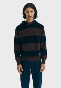 Jeune personne avec des cheveux bouclés portant un hoodie tricoté à rayures larges bleu et marron et un jean bleu foncé, se tenant devant un fond uni.