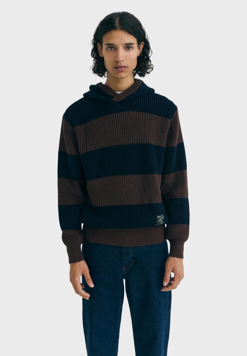 Jeune personne avec des cheveux bouclés portant un hoodie tricoté à rayures larges bleu et marron et un jean bleu foncé, se tenant devant un fond uni.