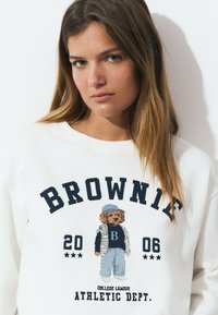 Sweatshirt blanc avec "BROWNIE" en bleu marine, illustré par un dessin d'un chien cartoon en tenue décontractée, accompagnée d'étoiles et de l'année "2006."