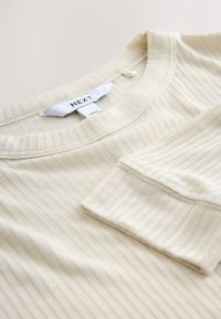 Cămașă bej deschis cu mâneci lungi, cu dungi, guler rotund, având un buzunar aplicat și o textură subtilă. Eticheta afișează "NEXT."