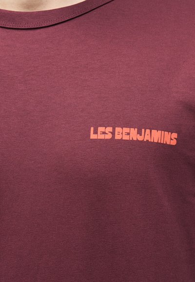Κοντινό πλάνο μιας μπορντό μπλούζας με το κείμενο "LES BENJAMINS" τυπωμένο πορτοκαλί στην πάνω αριστερή περιοχή του στήθους.