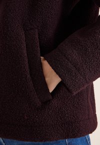 Una mano infilata nella tasca di una giacca in pile di un marrone scuro e texture, con del denim blu visibile sotto l'orlo della giacca.