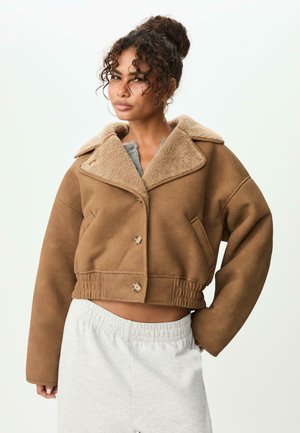 Afskåret brun jakke med krave af kunstpels, store knapper og sidelommer; sat sammen med lysegrå sweatsbukser. Blød tekstur.